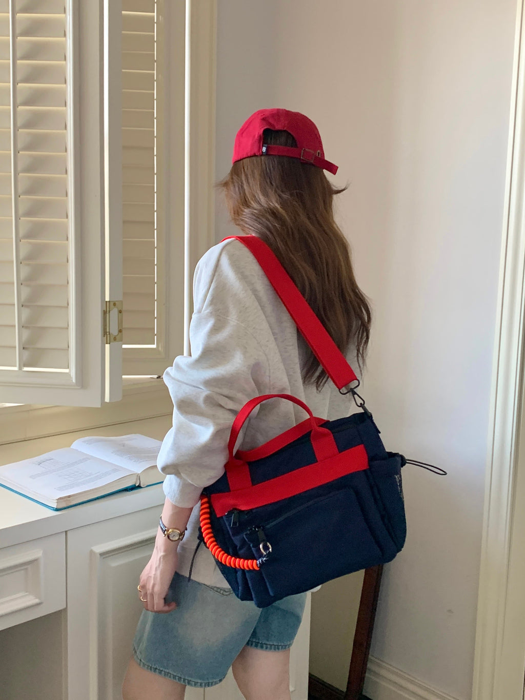 Person wearing a red cap and holding a navy and red bag in a room with a window and desk.