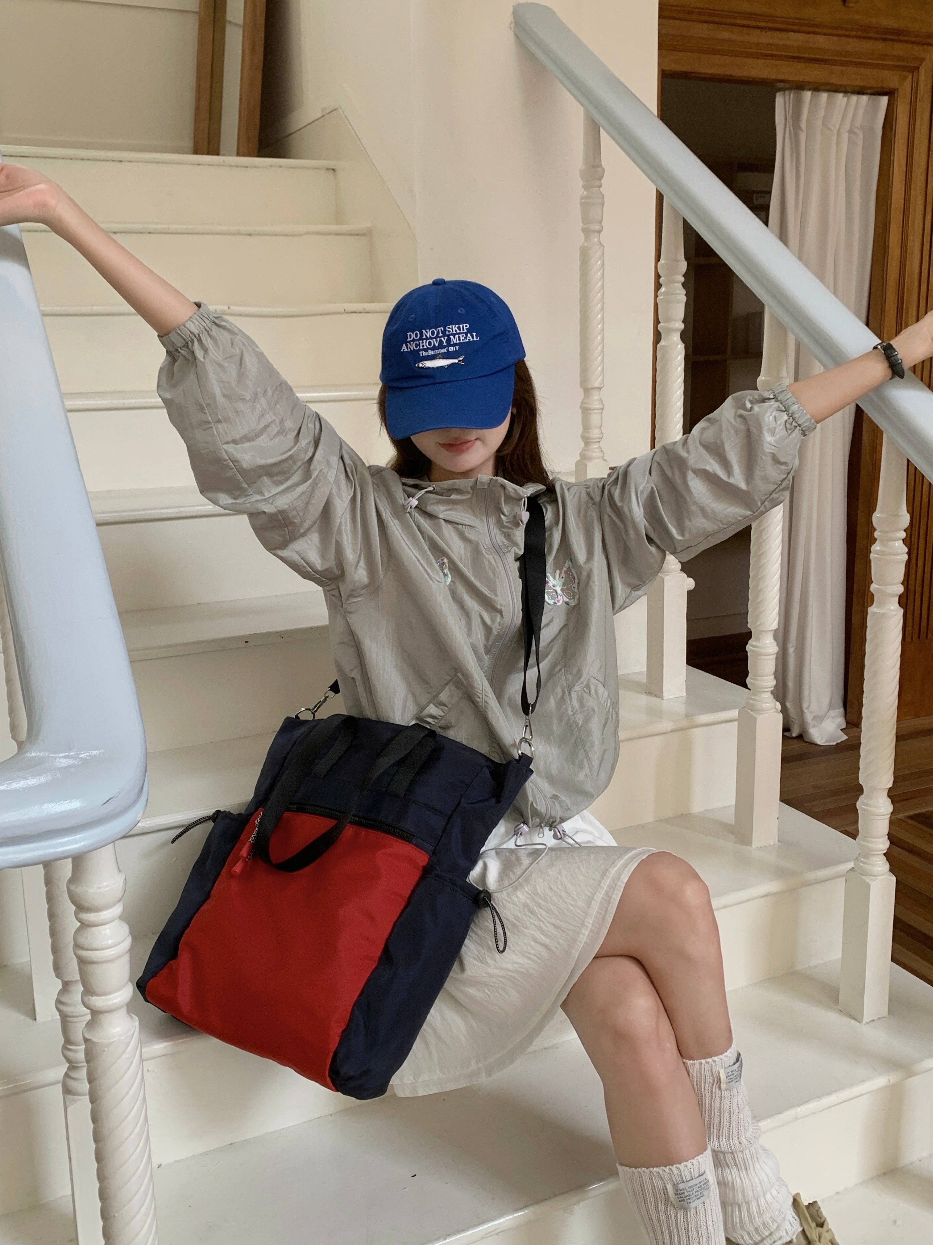 Person sitting on a staircase wearing a blue cap and light jacket with a red and navy bag.