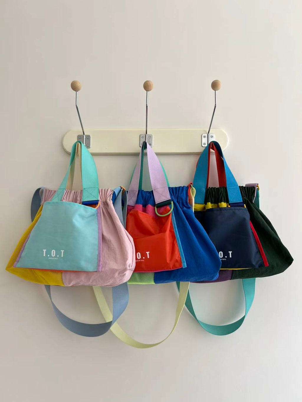 Four colorful T.O.T canvas tote bags in pastel and bright colors hanging from a cream-colored peg rail on a white wall.