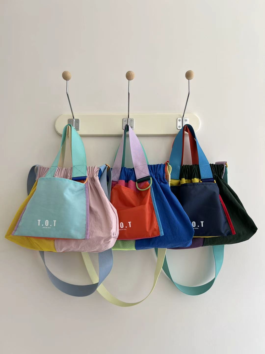 Four colorful T.O.T canvas tote bags in pastel and bright colors hanging from a cream-colored peg rail on a white wall.