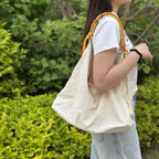 A person outdoors wearing a large off-white canvas shoulder bag with a colorful braided strap in orange, yellow, and green, set against a background of green foliage.