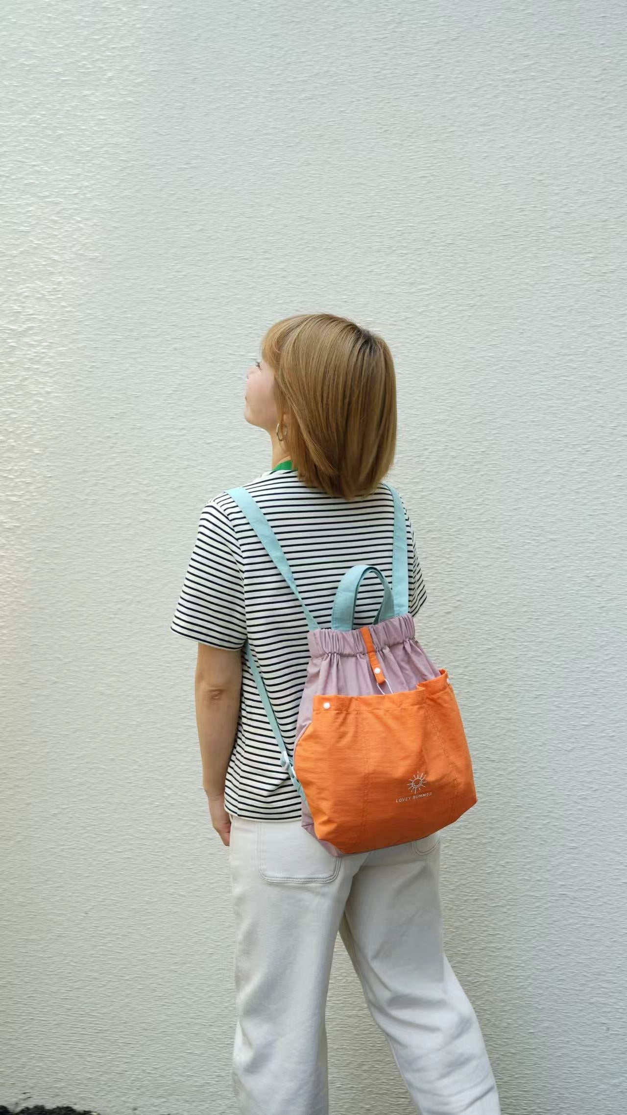 Person wearing a striped shirt and white pants with an orange backpack against a light gray wall.