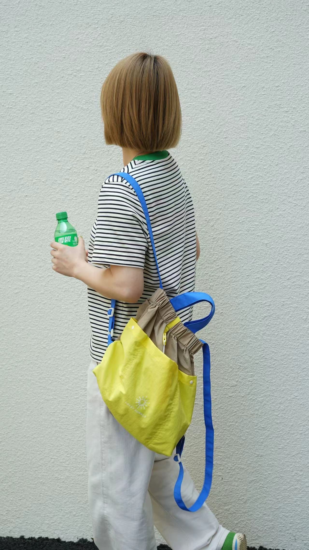 Person holding a yellow bag with a blue strap and a green bottle against a plain background