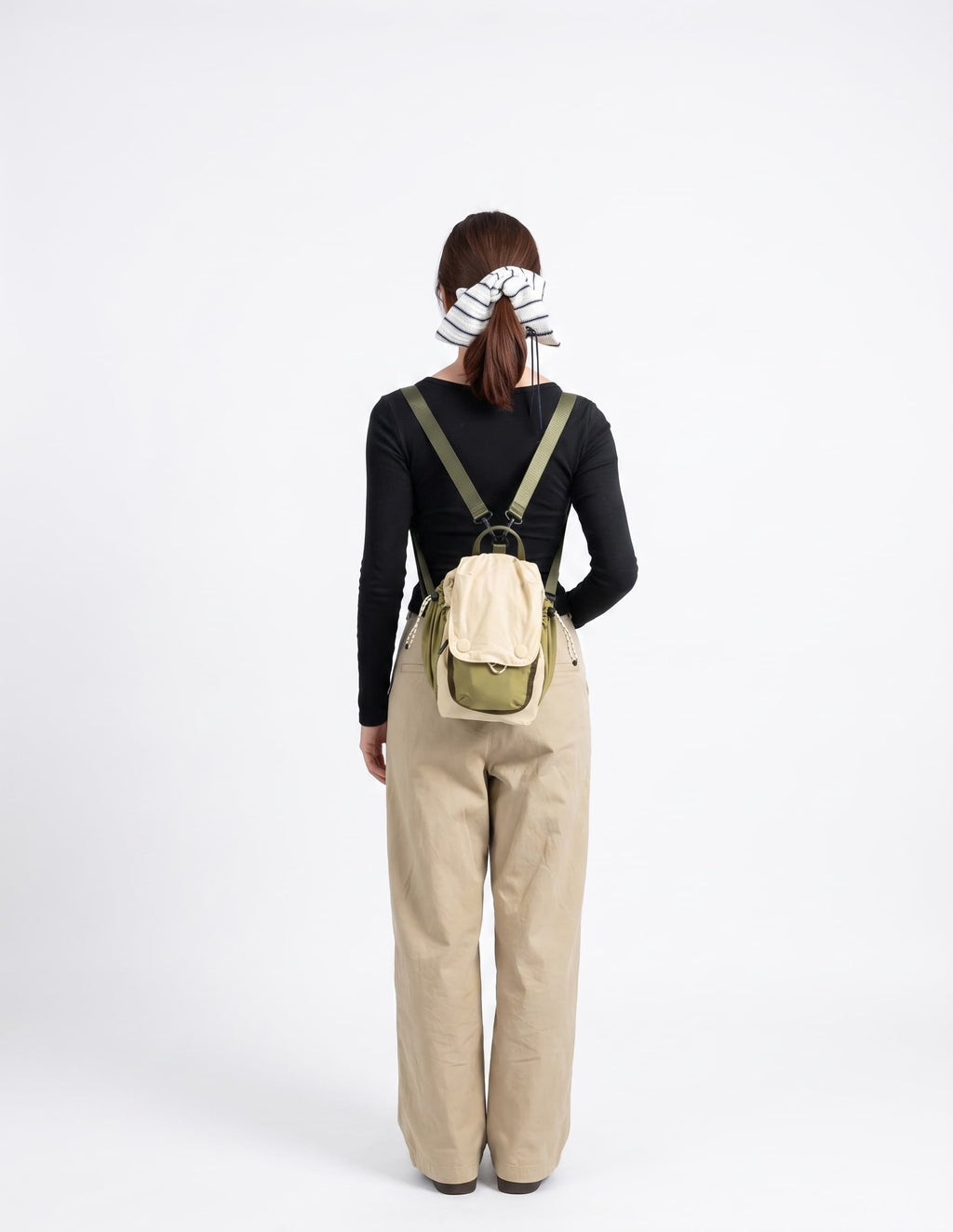 A woman viewed from behind wearing a small beige and olive green backpack, a black long-sleeve top, and beige wide-leg pants against a plain white studio background.
