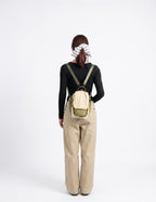 A woman viewed from behind wearing a small beige and olive green backpack, a black long-sleeve top, and beige wide-leg pants against a plain white studio background.