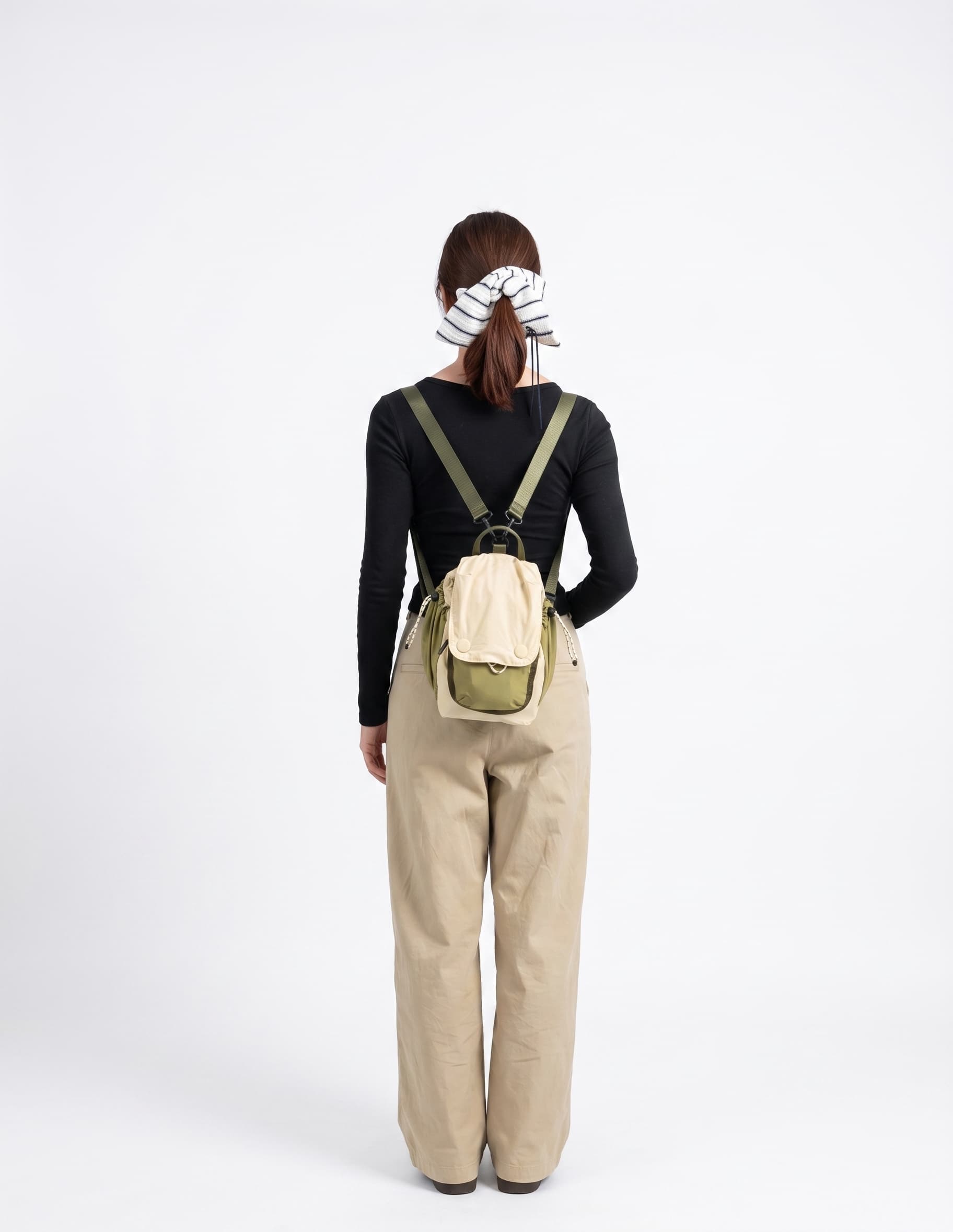 A woman viewed from behind wearing a small beige and olive green backpack, a black long-sleeve top, and beige wide-leg pants against a plain white studio background.
