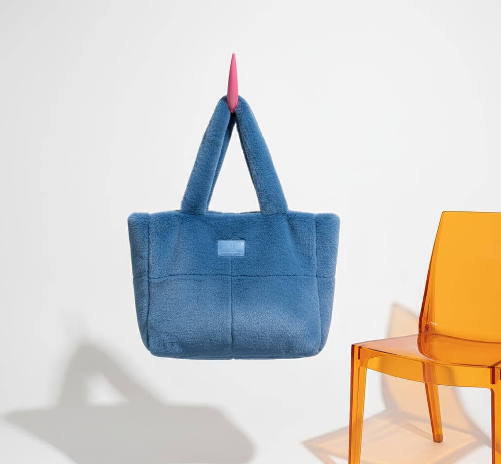 Blue tote bag with a small logo on a white background, next to an orange chair.Plush faux fur tote bag with padded handles, zippered top, and spacious interior – designed for cozy winter outings and everyday comfort. Soft, lightweight, and stylish, perfect for carrying essentials in cold weather. A chic and functional piece from Tales of Totes – standing out from Indian brands like Zouk, Chumbak, and Miraggio with unique textures and winter-ready fashion flair."
