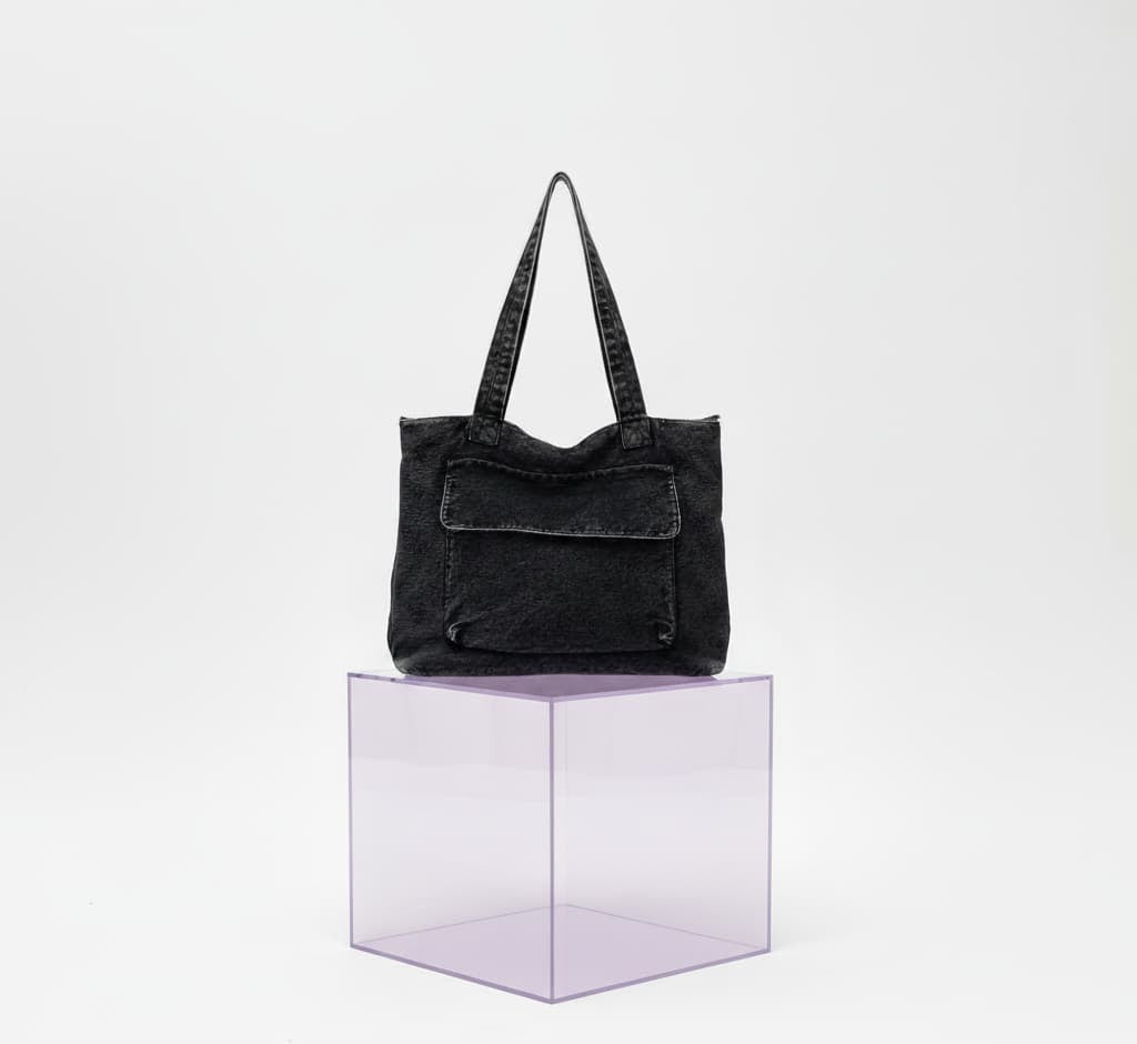 Black denim tote bag on a clear cube with a white background."Stylish denim tote bags with front flap pockets and twin shoulder straps – perfect for casual outings, college, or everyday errands. Made from durable washed denim for a rugged streetwear look with spacious interior and secure zip closure. A versatile and functional carryall from Tales of Totes – redefining everyday fashion bags in India beyond Zouk, DailyObjects, and Chumbak."
