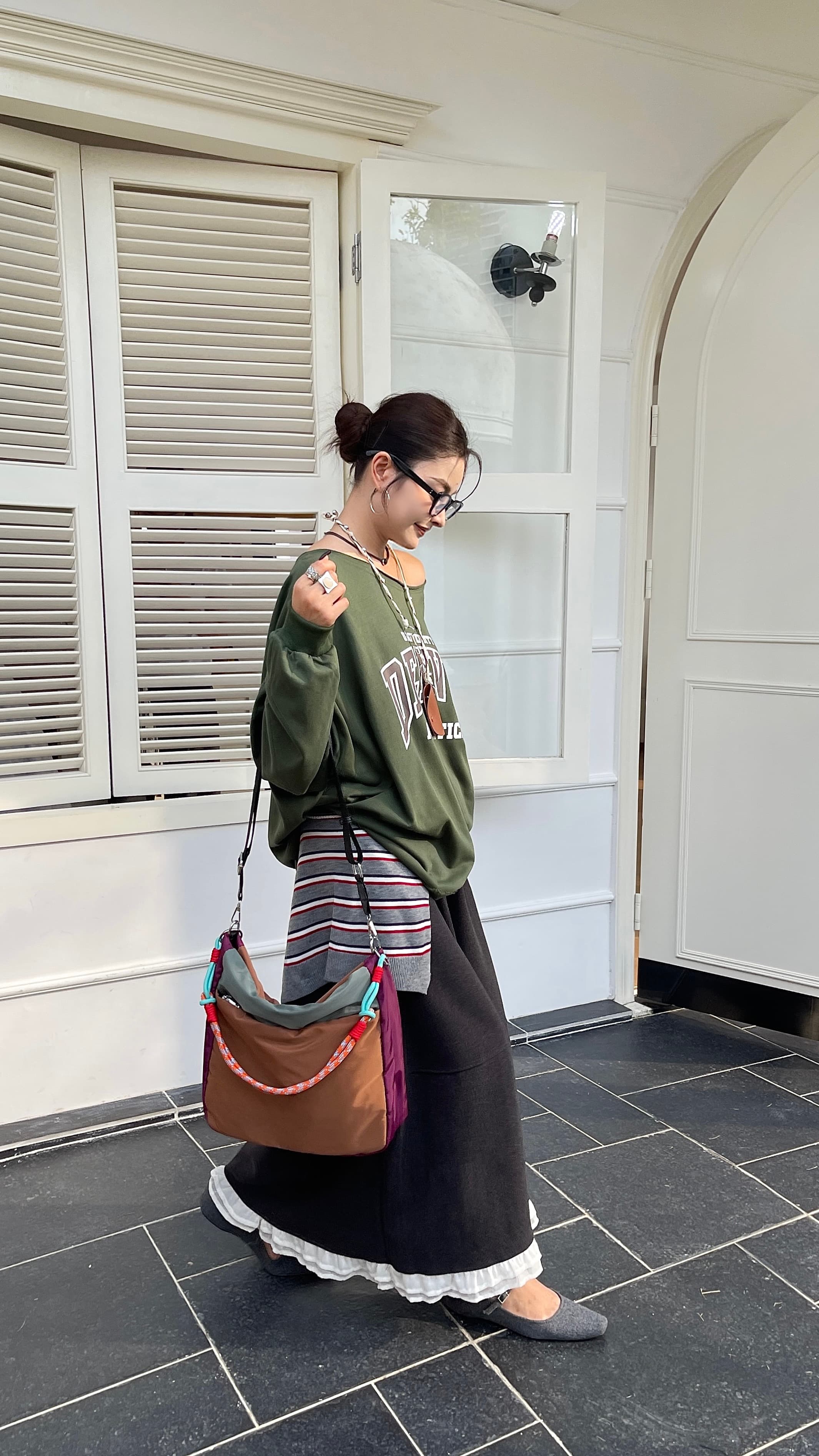 Woman standing outside a building, wearing a green sweatshirt and dark pants, with a colorful bag. Multi-pocket travel tote bag with 20-litre capacity and compact dimensions (35 x 35 x 10 cm), featuring a wide adjustable strap and color-blocked utility design. Lightweight, durable, and ideal for travel, college, or daily use. A high-performance everyday essential from Tales of Totes – built for comfort, style, and smart organization in India’s fast-paced lifestyle."
