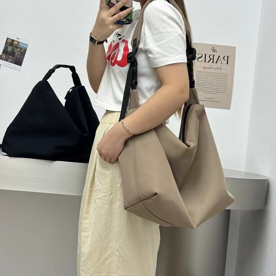 Person holding a beige handbag with a black bag and white wall in the background.Convertible nylon tote bag with detachable crossbody strap, zippered front pocket, and main compartment. Available in olive green, black, and beige.