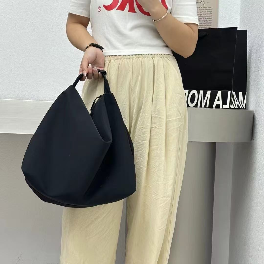 Person holding a black bag wearing a white t-shirt with red text and beige pants.Convertible nylon tote bag with detachable crossbody strap, zippered front pocket, and main compartment. Available in olive green, black, and beige.