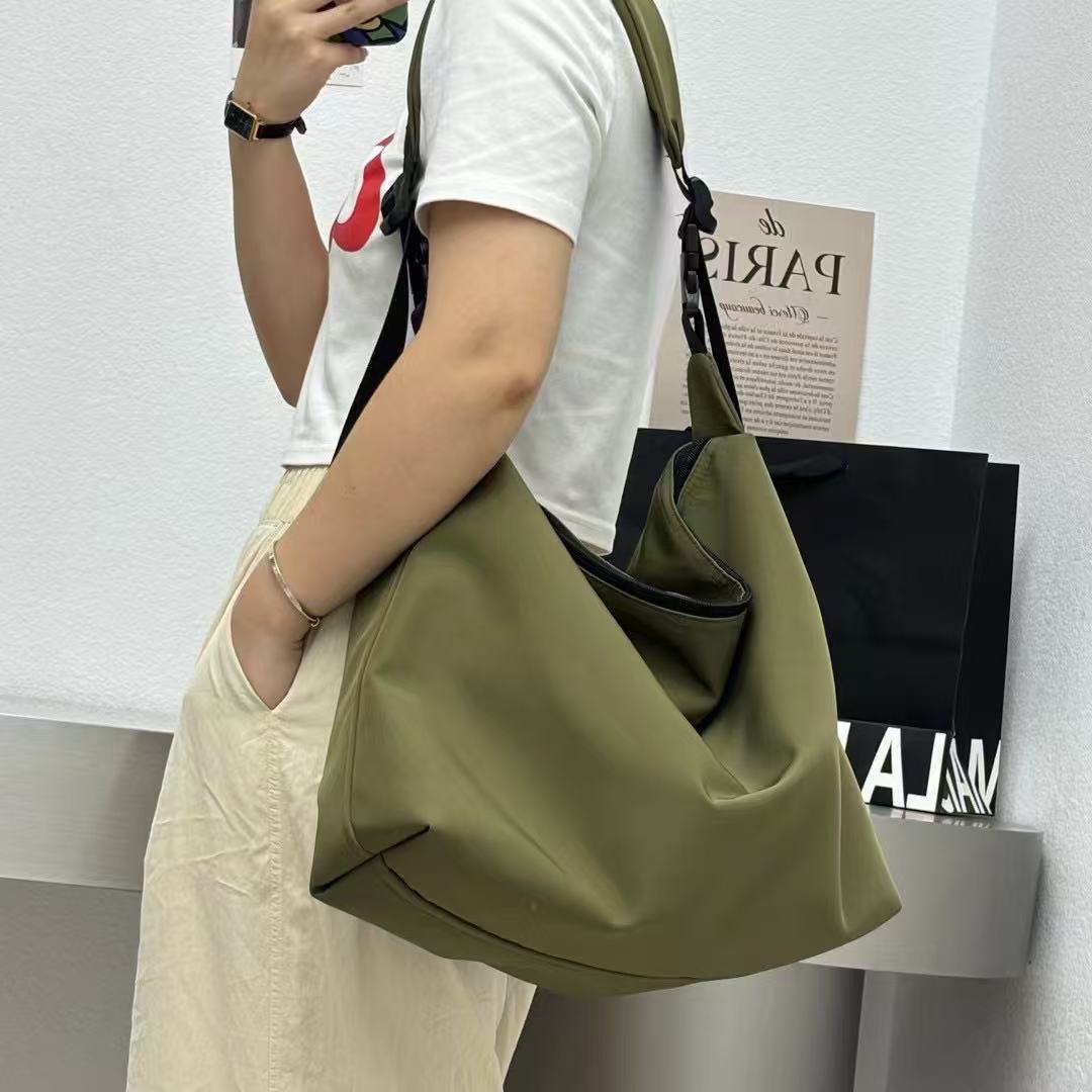 Person holding a green handbag with a white wall and black bag in the background.Convertible nylon tote bag with detachable crossbody strap, zippered front pocket, and main compartment. Available in olive green, black, and beige.