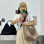 Person holding a beige bag, wearing a white t-shirt with red text and a beige cap, taking a mirror selfie. Convertible nylon tote bag with detachable crossbody strap, zippered front pocket, and main compartment. Available in olive green, black, and beige.