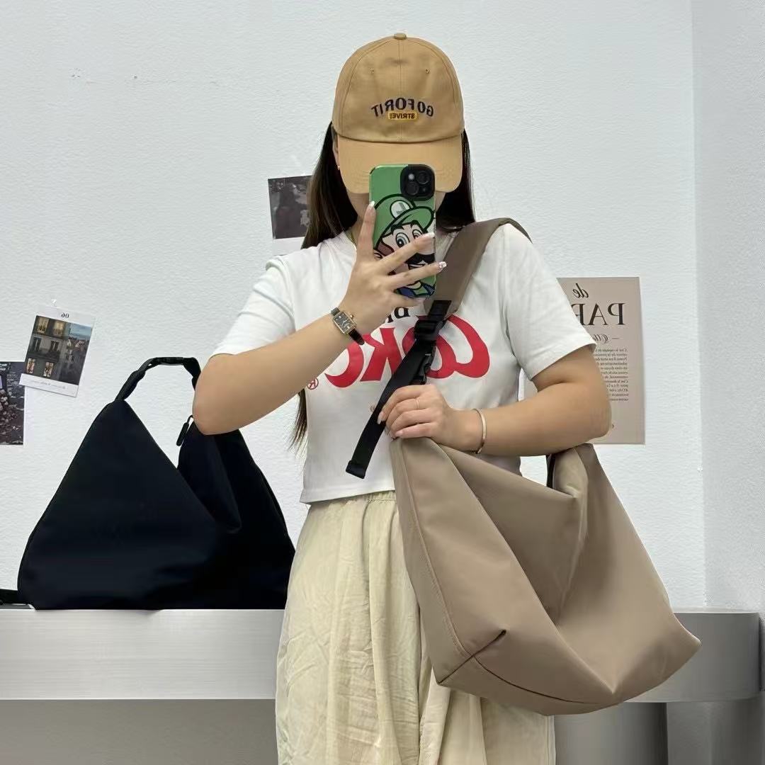 Person holding a beige bag, wearing a white t-shirt with red text and a beige cap, taking a mirror selfie. Convertible nylon tote bag with detachable crossbody strap, zippered front pocket, and main compartment. Available in olive green, black, and beige.