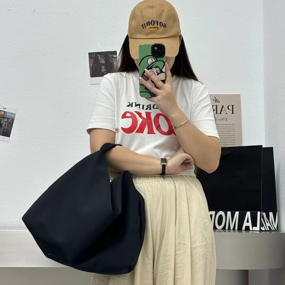 Person wearing a white t-shirt with red text, beige cap, and holding a black bag.Convertible nylon tote bag with detachable crossbody strap, zippered front pocket, and main compartment. Available in olive green, black, and beige.