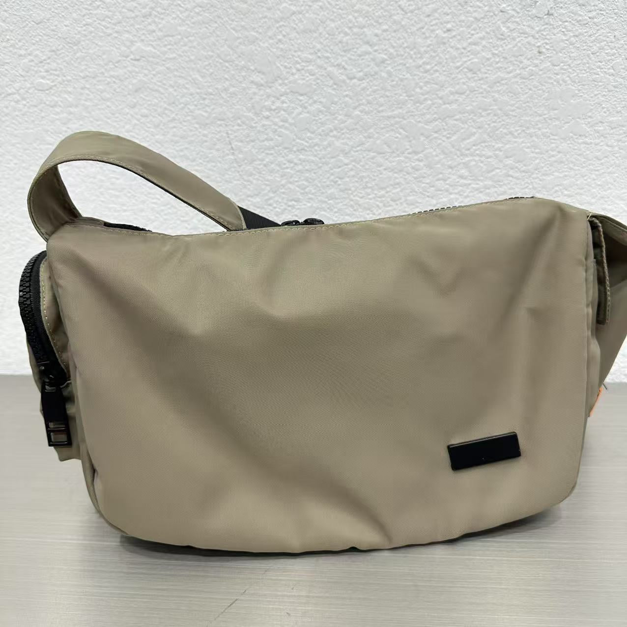 Beige shoulder bag on a light gray surface with a white textured backgroundMini utility tote bag 33x22x14 cm with multiple pockets, zip closure, and water-resistant nylon material. Everyday carry tote for men and women, ideal for office, travel, and casual use in India. Alternative to Muji tote, Decathlon utility bag, and Wildcraft crossbody. Features bright orange inner lining and durable adjustable strap.