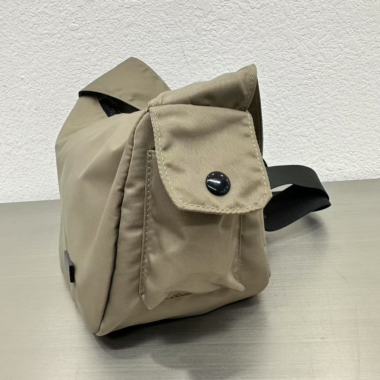 Beige bag with a black button on a light gray surface and white textured background,Mini utility tote bag 33x22x14 cm with multiple pockets, zip closure, and water-resistant nylon material. Everyday carry tote for men and women, ideal for office, travel, and casual use in India. Alternative to Muji tote, Decathlon utility bag, and Wildcraft crossbody. Features bright orange inner lining and durable adjustable strap.