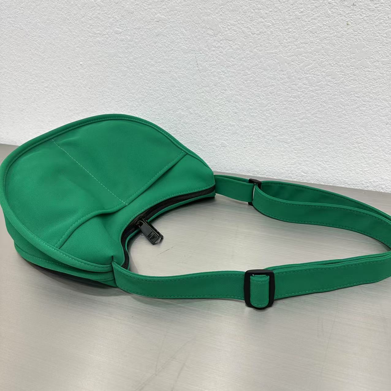 Green bag with a strap on a light gray surface,Trendy emerald green crescent half-moon shoulder bag for women, durable canvas, versatile for daily use, ethnic wear, and casual outings in India. This stylish sling bag offers a modern minimalist design, spacious interior, functioning as a chic carryall or small hobo tote.