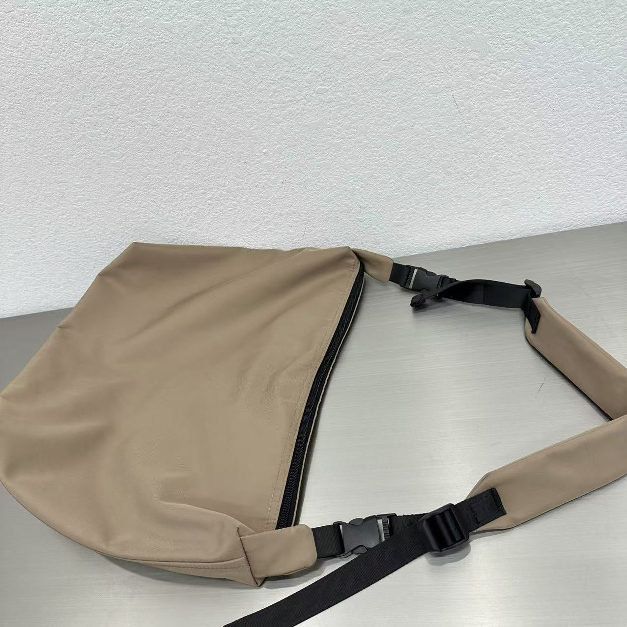 Beige sling bag with black straps on a light gray surface,Convertible nylon tote bag with detachable crossbody strap, zippered front pocket, and main compartment. Available in olive green, black, and beige.