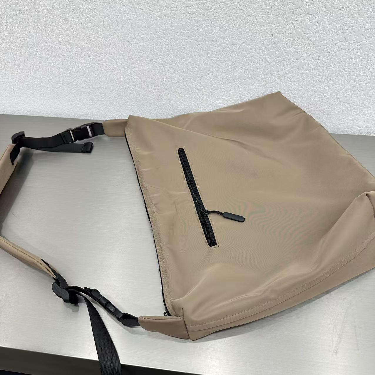 Beige bag with a black strap on a light gray surface,Convertible nylon tote bag with detachable crossbody strap, zippered front pocket, and main compartment. Available in olive green, black, and beige.