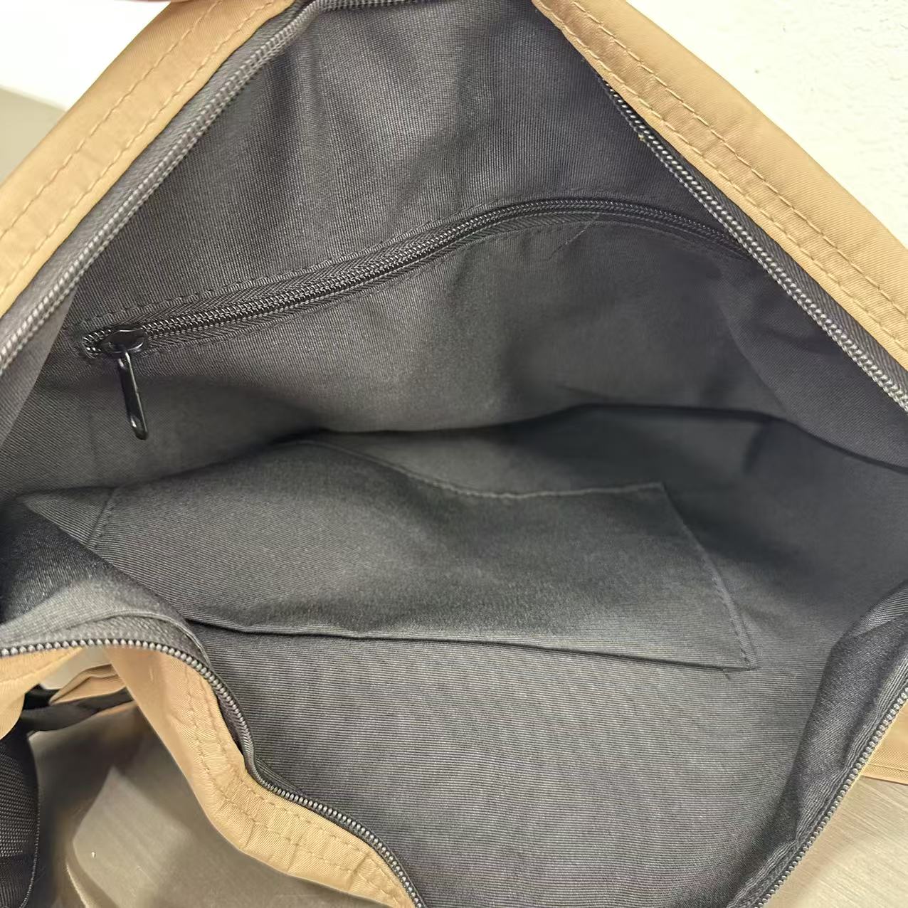 Open beige bag with black interior lining on a light wooden surface,Convertible nylon tote bag with detachable crossbody strap, zippered front pocket, and main compartment. Available in olive green, black, and beige.