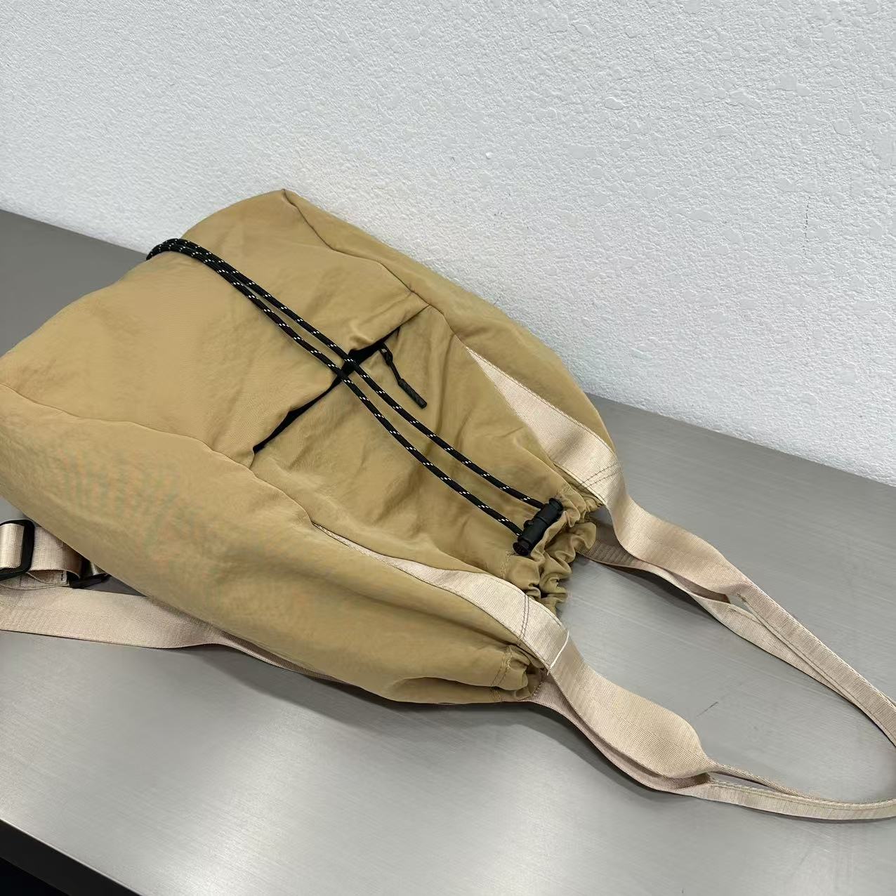 Beige bag with black straps on a gray surface."Stylish khaki drawstring tote bag with multiple compartments and adjustable straps, perfect for daily use, shopping, and travel in India. Features include a secure zippered pocket, spacious main compartment, and comfortable shoulder straps. Ideal for fashion-conscious individuals seeking a versatile and trendy bag."