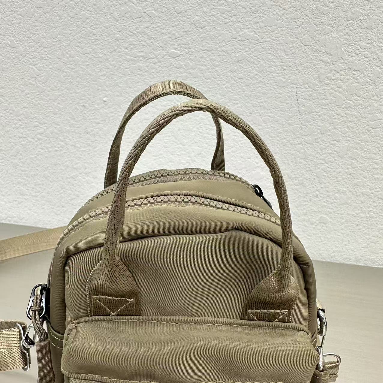 Khaki backpack with multiple straps on a textured light gray background,A high-fashion shot of a woman in a black skirt and white shirt, holding a stylish, minimalist brown bag. The image is professionally lit in a studio against a clean white background, with a strong focus on the bag's sleek design and texture. The photo emphasizes a sophisticated and modern aesthetic, perfect for luxury or premium lifestyle brands.