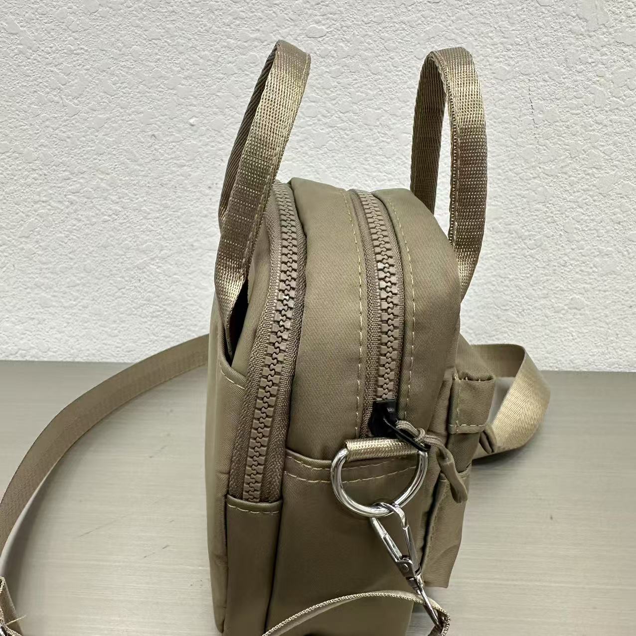 Beige backpack with a zipper on a light gray surface,A high-fashion shot of a woman in a black skirt and white shirt, holding a stylish, minimalist brown bag. The image is professionally lit in a studio against a clean white background, with a strong focus on the bag's sleek design and texture. The photo emphasizes a sophisticated and modern aesthetic, perfect for luxury or premium lifestyle brands.