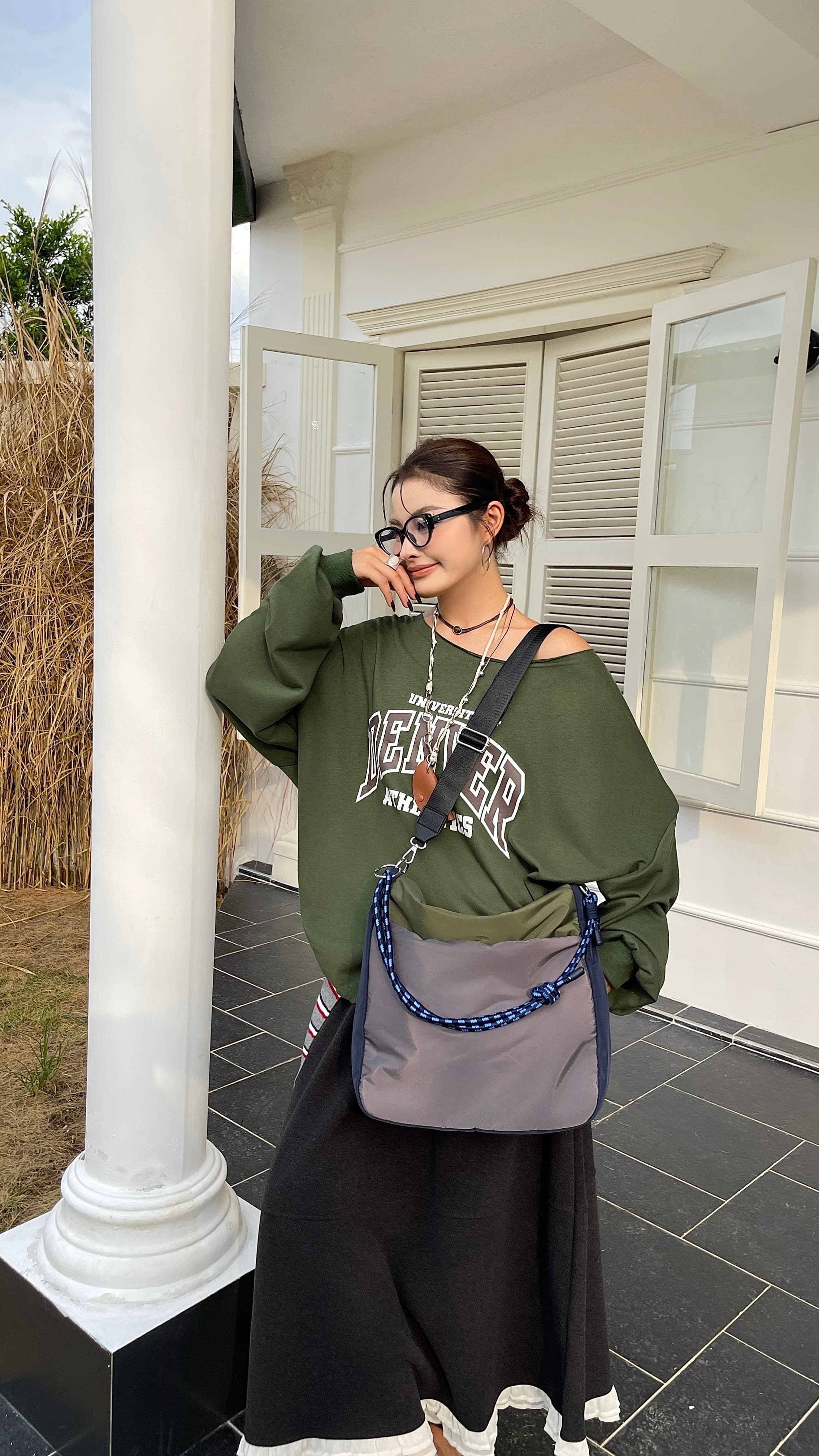 Woman wearing a green sweatshirt and black skirt with a gray and blue bag, standing on a porch. Multi-pocket travel tote bag with 20-litre capacity and compact dimensions (35 x 35 x 10 cm), featuring a wide adjustable strap and color-blocked utility design. Lightweight, durable, and ideal for travel, college, or daily use. A high-performance everyday essential from Tales of Totes – built for comfort, style, and smart organization in India’s fast-paced lifestyle."
