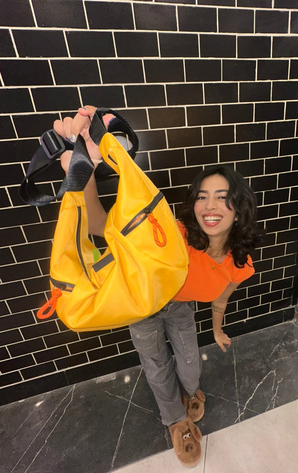 Person holding a yellow bag against a black tiled wall,Collage of a black and orange bag with different angles showing its design and features.Waterproof tote bag with a durable exterior and a bright orange interior. Features an adjustable strap and multiple pockets for organization. Ideal for travel, gym, or daily use in India.