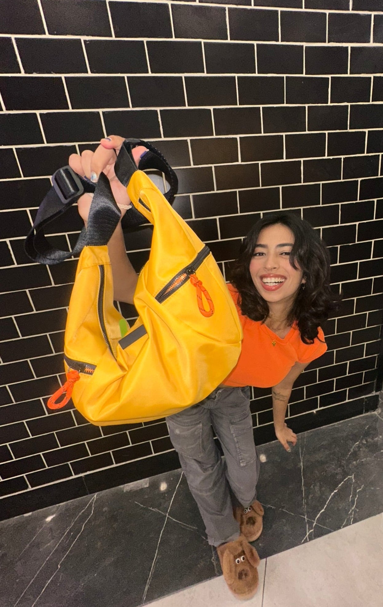 Person holding a yellow bag against a black tiled wall,Collage of a black and orange bag with different angles showing its design and features.Waterproof tote bag with a durable exterior and a bright orange interior. Features an adjustable strap and multiple pockets for organization. Ideal for travel, gym, or daily use in India.
