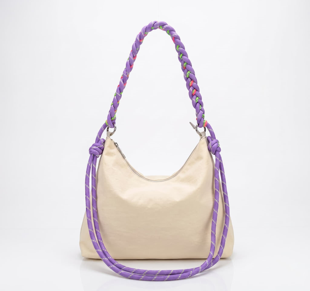 A cream-colored canvas hobo bag with a thick, braided purple shoulder strap featuring multicolor threads, photographed on a white background.