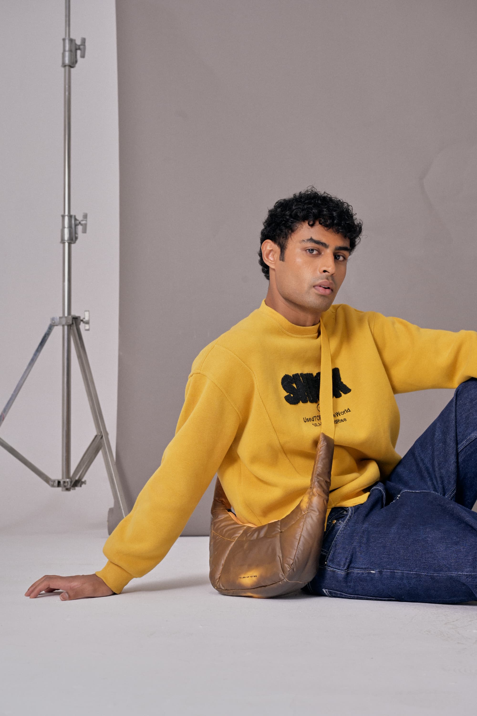 Man wearing a yellow sweatshirt with text, sitting on the floor against a plain background