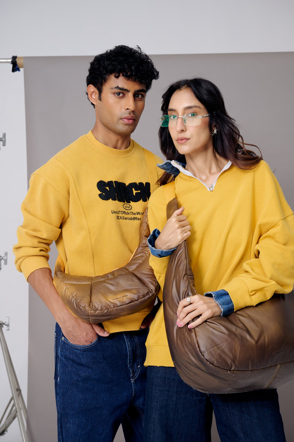 Two people wearing yellow sweatshirts with brown bags against a plain background