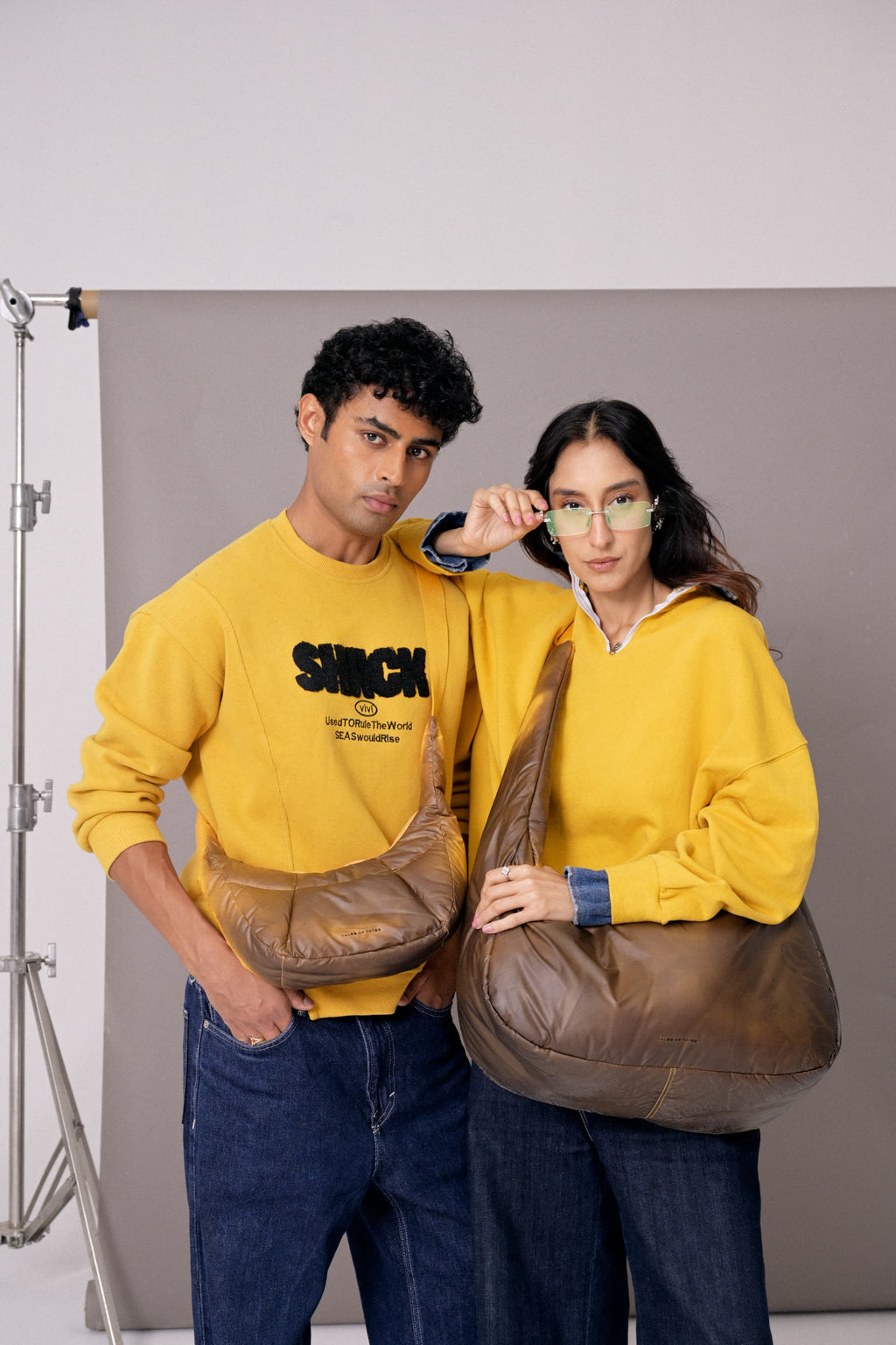 Two people wearing yellow sweatshirts holding brown leather bags against a plain background
