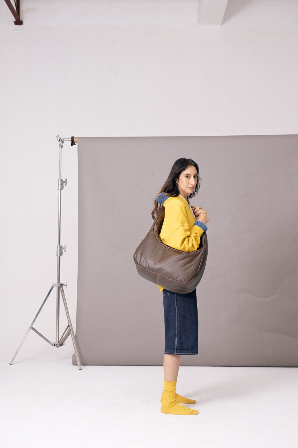 Woman holding a large brown bag in front of a gray backdrop with photography equipment.