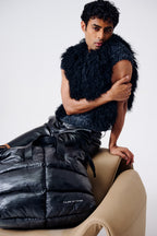 Man wearing a black fur vest and puffer jacket sitting on a beige chair against a white background