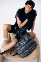 Man sitting on a chair with a large black backpack, wearing a dark fur vest and black pants.