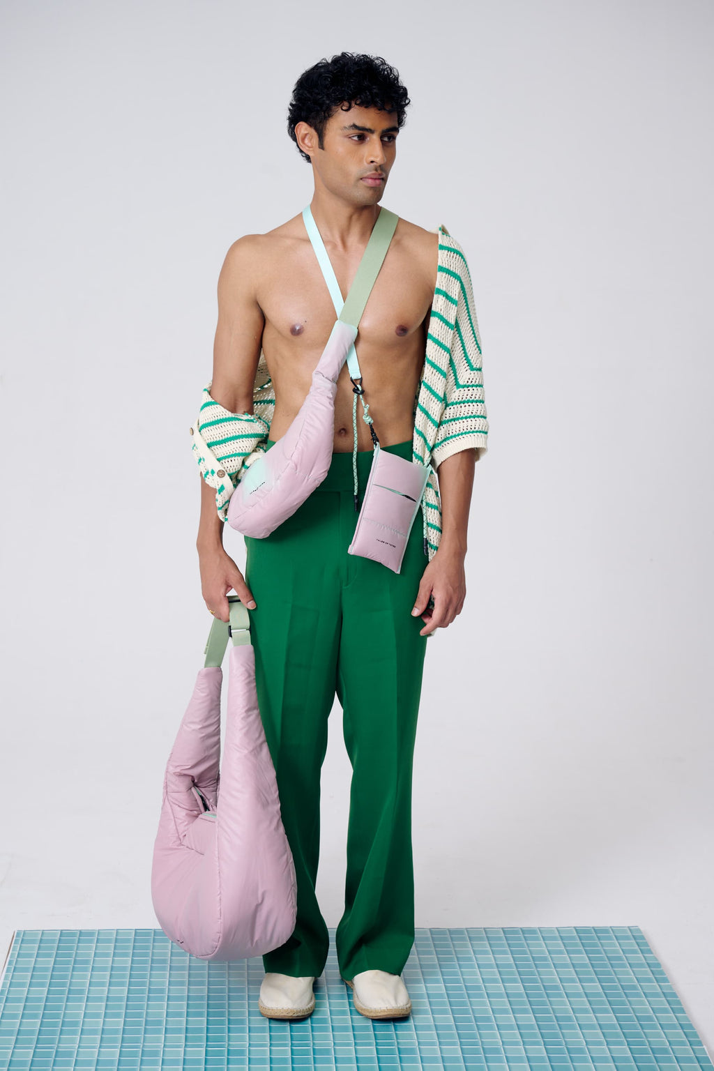 Man wearing a green and white striped shirt and green pants, holding pink bags on a grid floor.