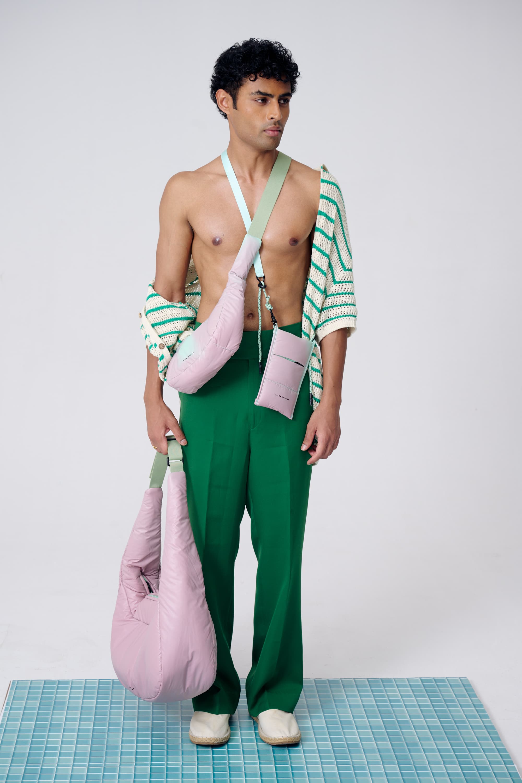 Man wearing a green and white striped shirt and green pants, holding pink bags on a grid floor.