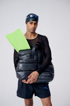Man holding a black pack with a green folder, wearing a black outfit and headband.