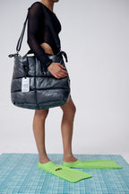Person holding a large black puffer bag on a tiled floor with green shoes.