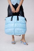 Person holding a large light blue puffer bag with black straps on a plain background